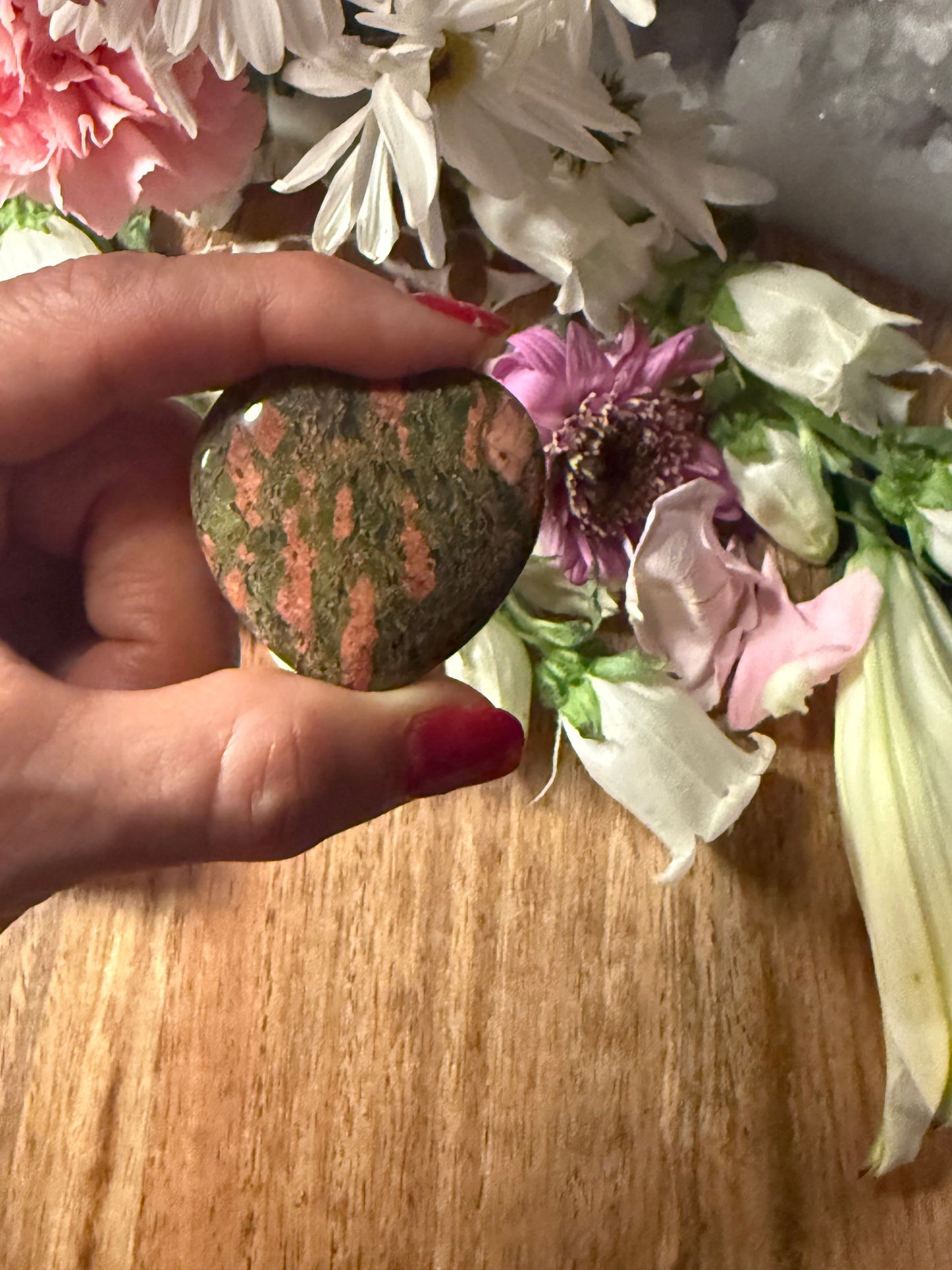 Hand holding a Unakite Crystal Heart stone with flowers in the background