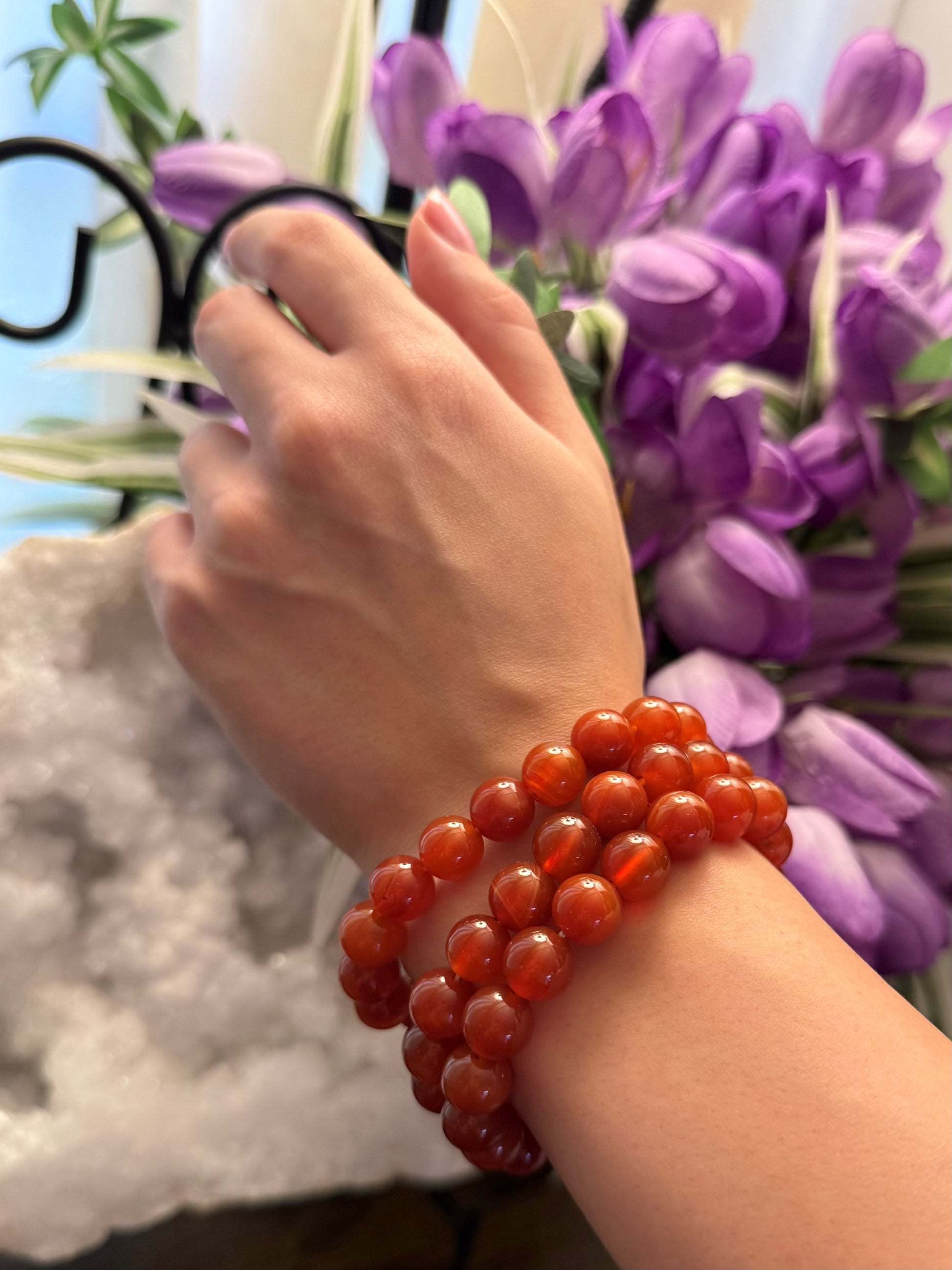 Hand wearing a red Carnelian beaded bracelet with a blurred background of purple flowers