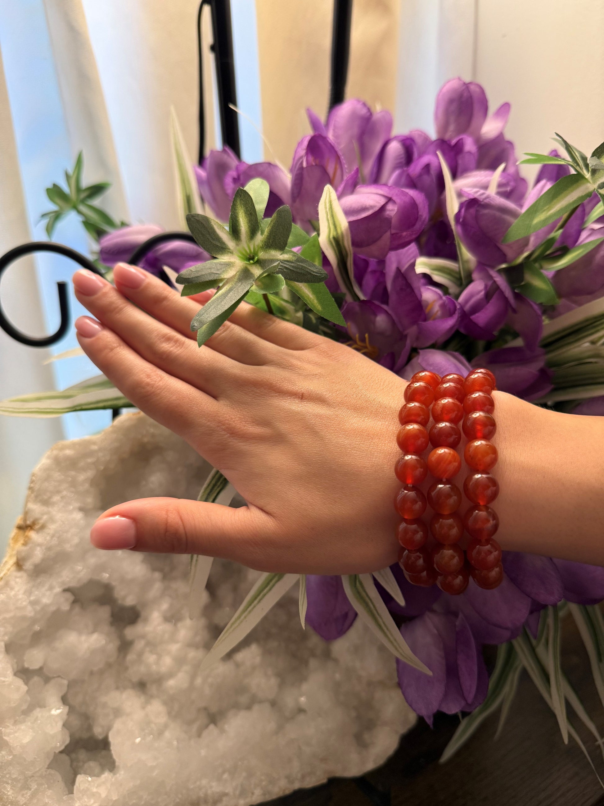 Hand wearing red Carnelian beaded bracelets holding purple flowers with a blurred background