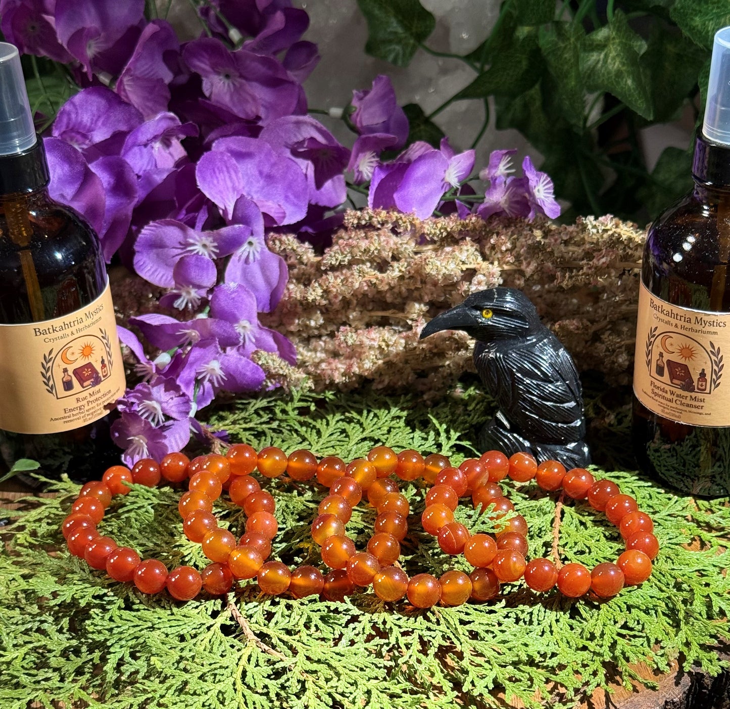 Two amber Carnelian bracelets on a green surface with purple flowers and a black raven figurine in the background.