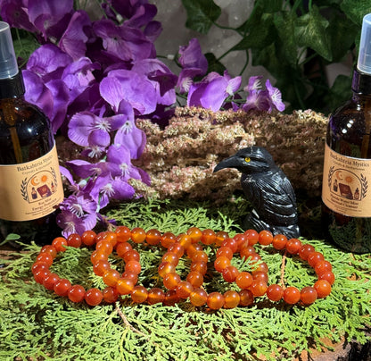 Two amber Carnelian bracelets on a green surface with purple flowers and a black raven figurine in the background.