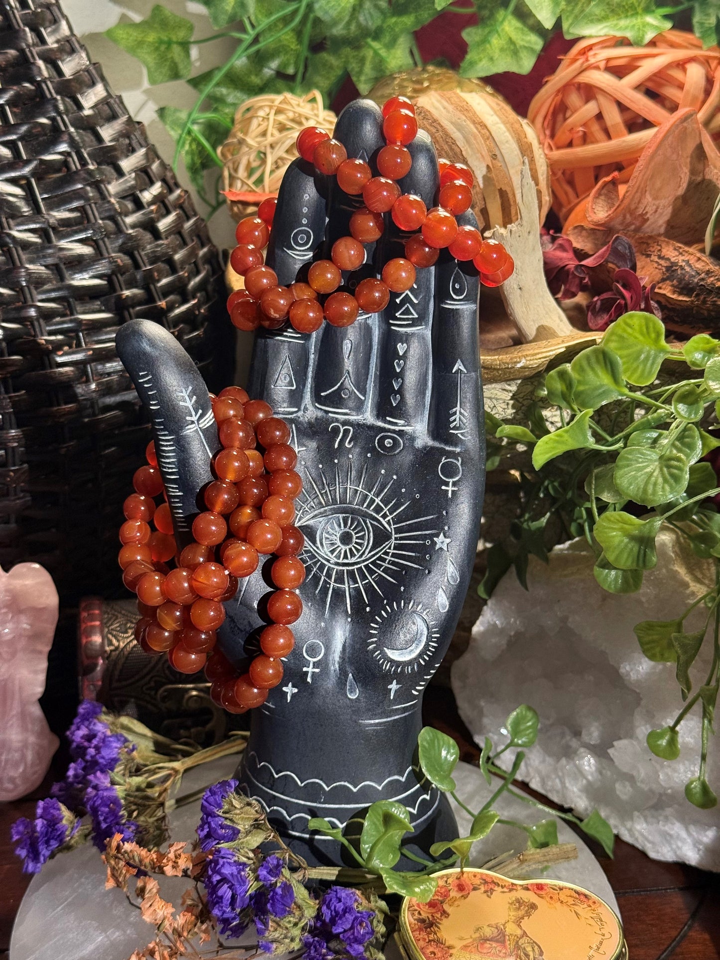 Decorative hand sculpture with red Carnelian bead bracelets on a mystical background with plants and crystals.