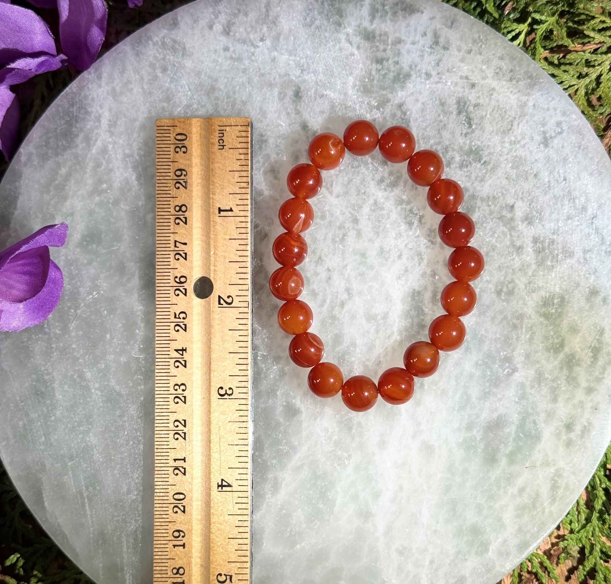 Red beaded Carnelian bracelet next to a ruler on a textured surface with purple flowers.
