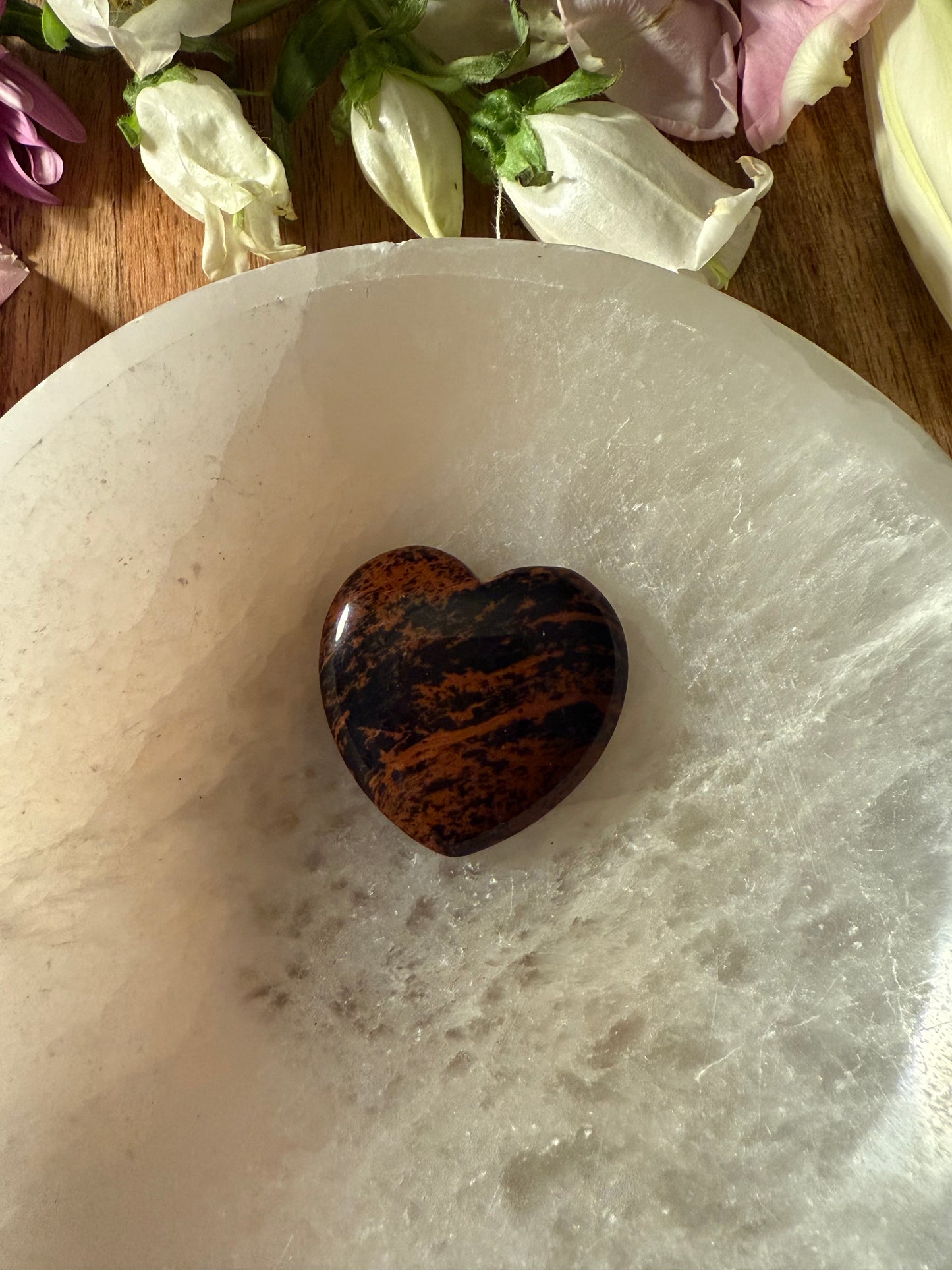 Mahogany Obsidian Crystal Heart stone on a white marble surface with flowers in the background