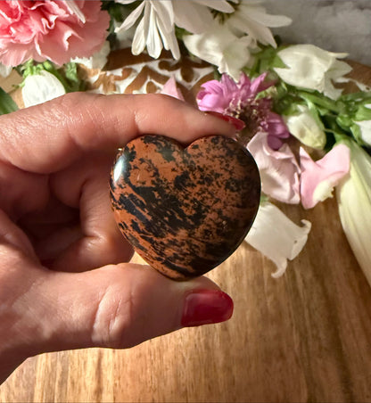 Hand holding a Mahogany Obsidian Crystal Heart stone with flowers in the background