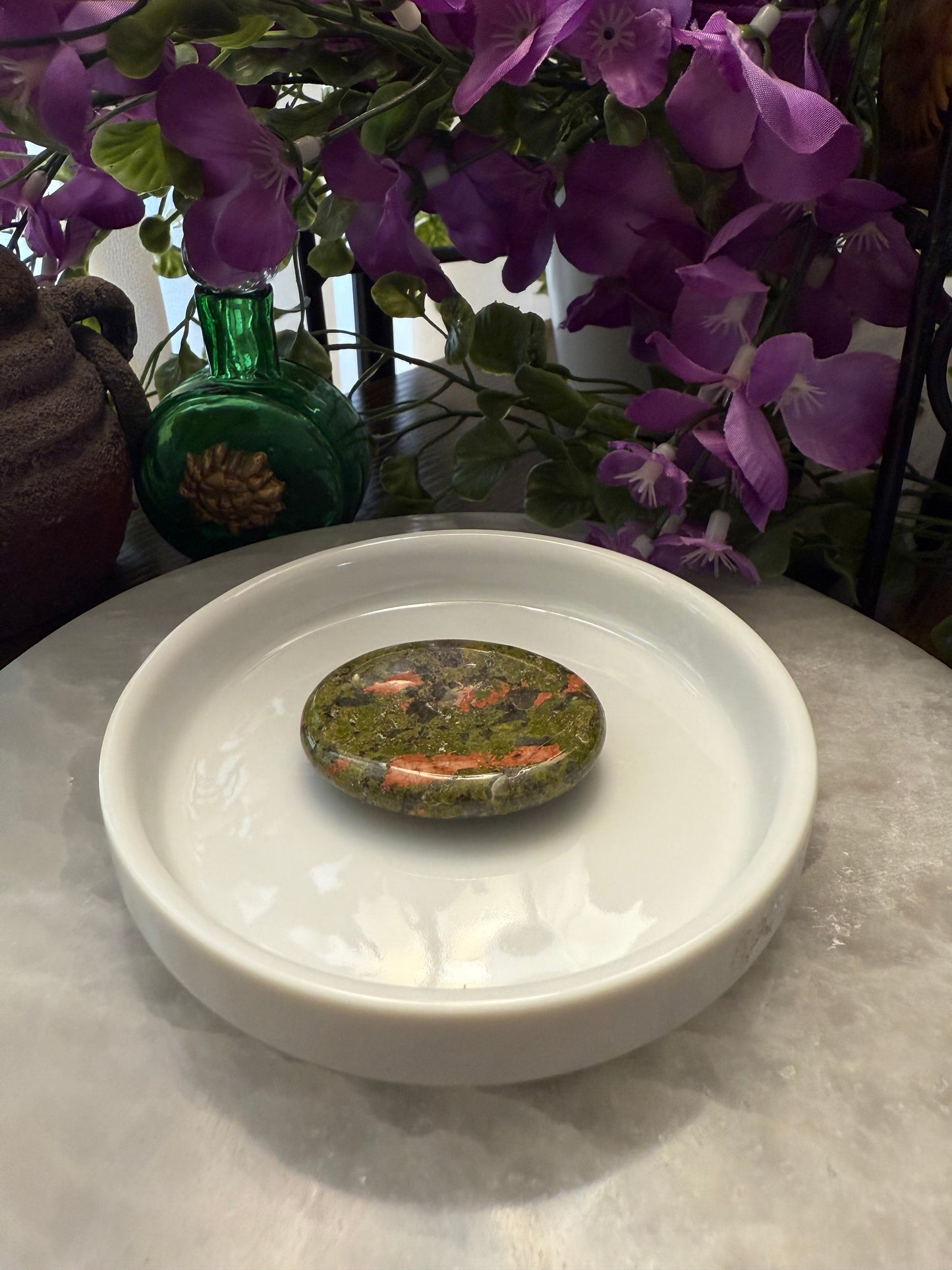 Round stone with natural patterns on a white plate, surrounded by purple flowers and a green bottle.