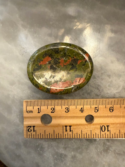 Oval-shaped stone with green, red, and black colors on a gray surface with a wooden ruler for scale.