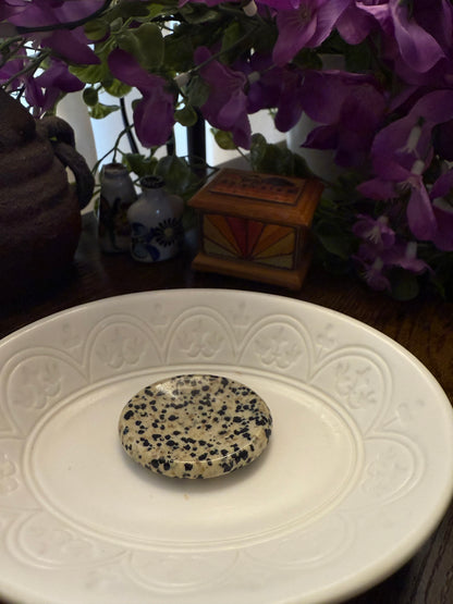 Dalmatian Jasper Worry Calming Stone on a white plate with decorative items and purple flowers in the background
