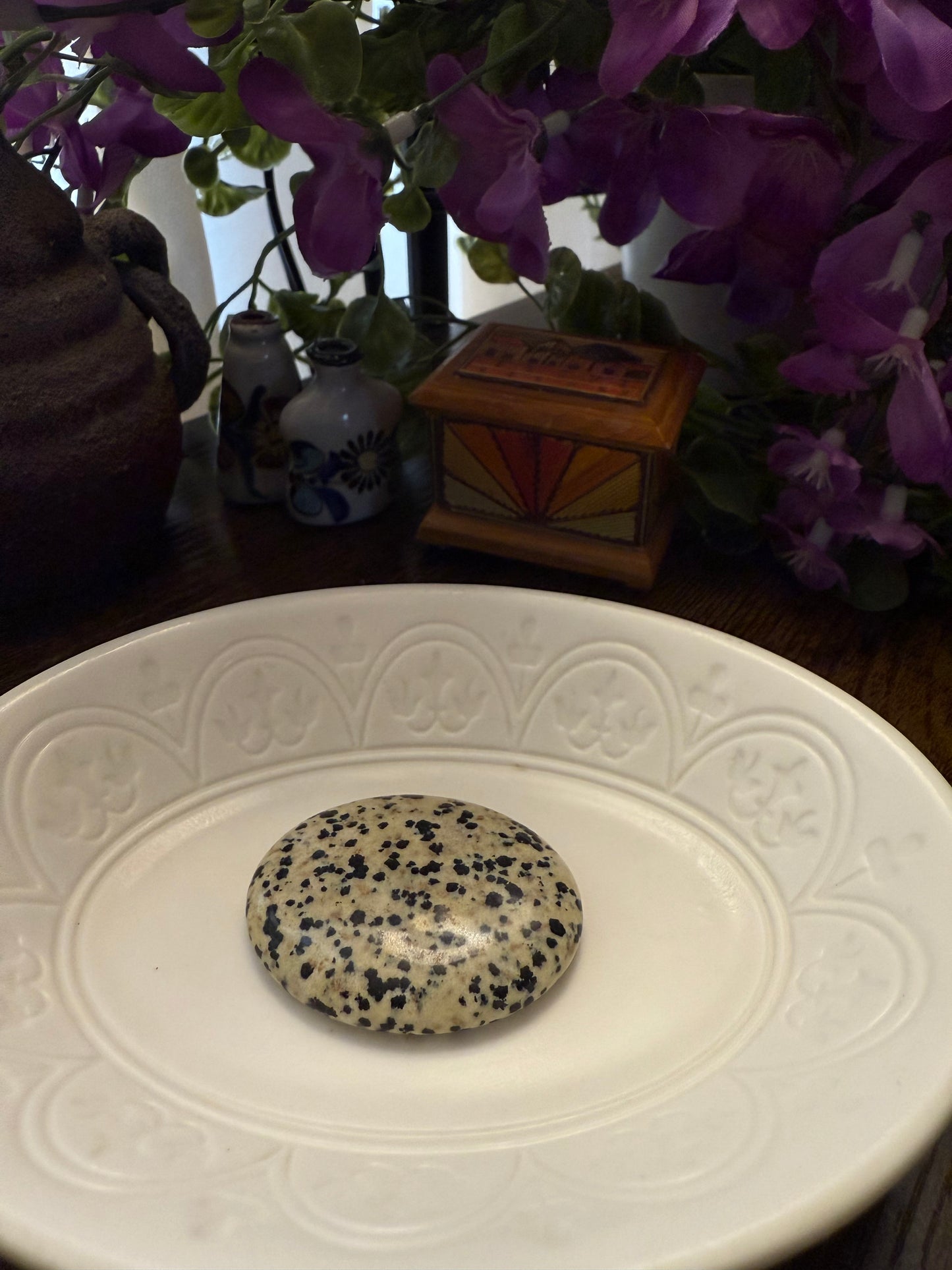 Dalmatian Jasper Worry Calming Stone on a white plate with a blurred background of flowers and decorative items.