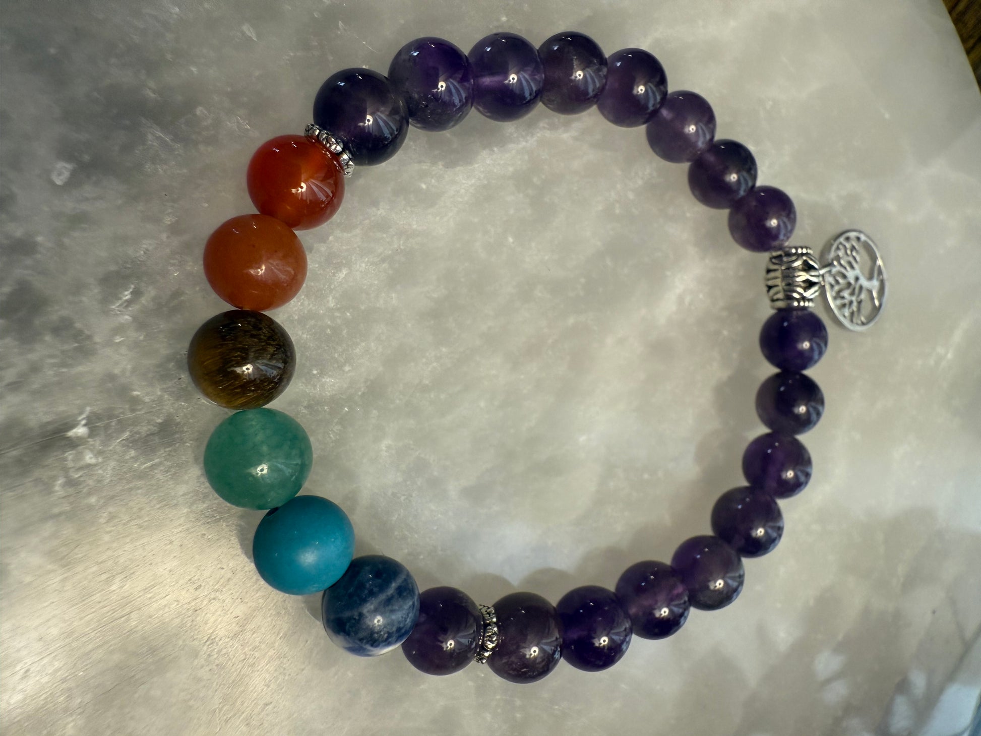 Multicolored Amethyst Chakra Balance Bracelet with a silver tree of life charm on a textured surface
