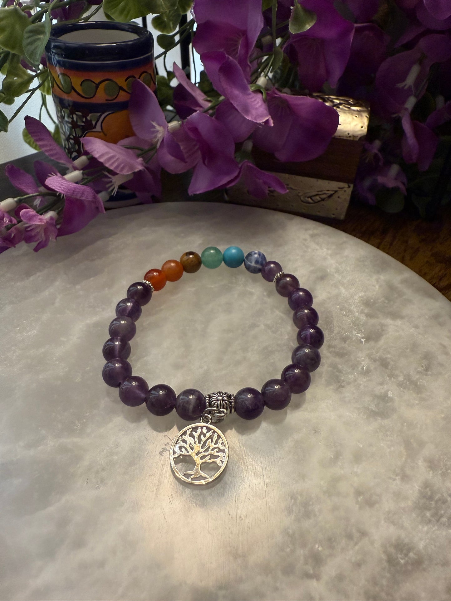 Amethyst Chakra Balance Bracelet with a tree of life charm on a textured surface with purple flowers in the background