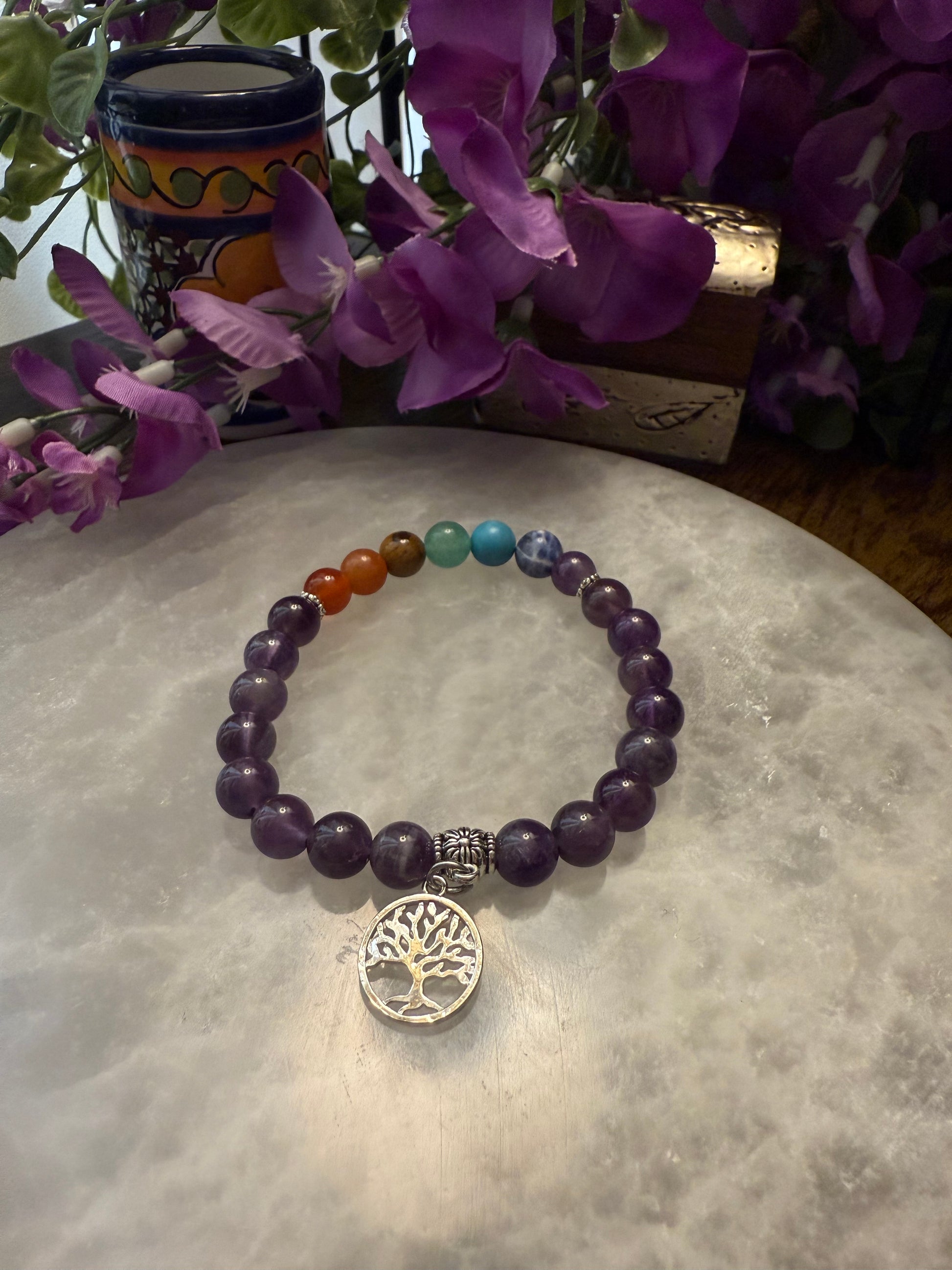 Amethyst Chakra Balance Bracelet with a tree of life charm on a textured surface with purple flowers in the background