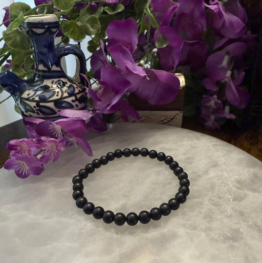 Black Natural Shungite Bead Bracelet on a white surface with purple flowers and a decorative vase in the background.