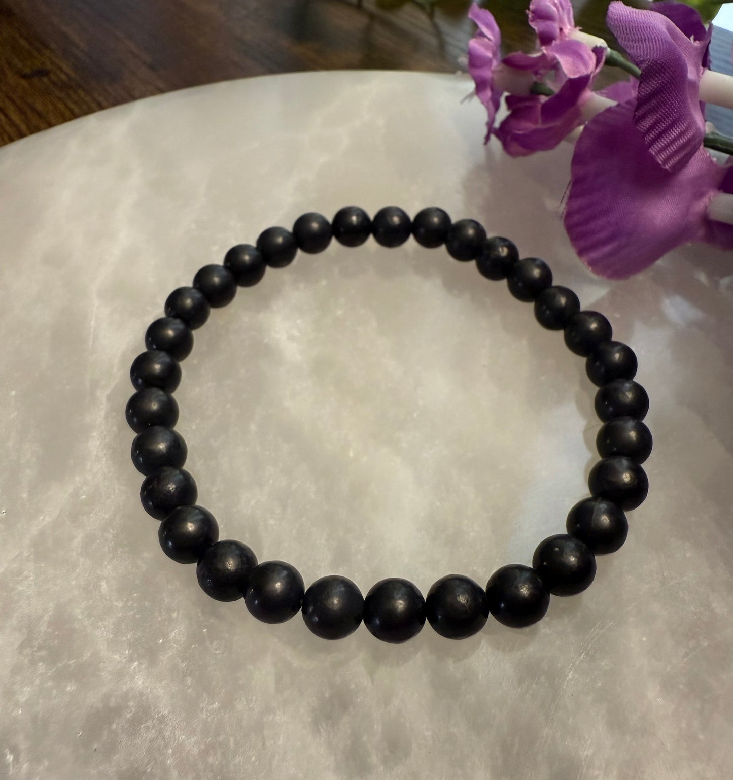 Black Natural Shungite Bead Bracelet on a light surface with purple flowers in the background