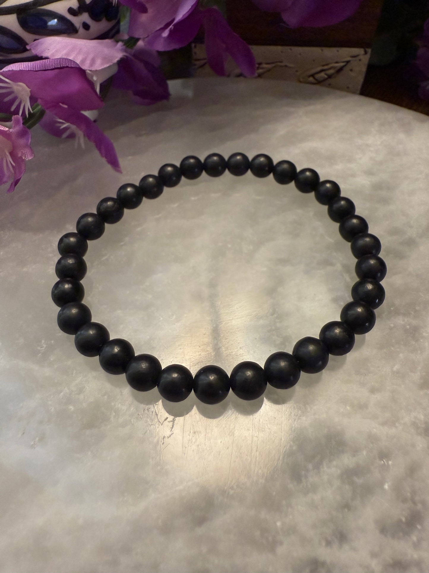 Black Natural Shungite Bead Bracelet on a textured surface with purple flowers in the background