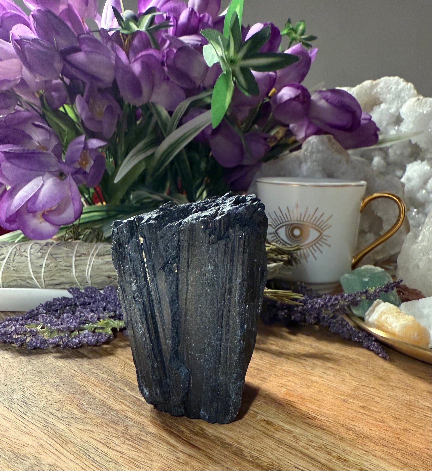 Decorative setting with a black crystal, purple flowers, and a cup with an eye design on a wooden surface.