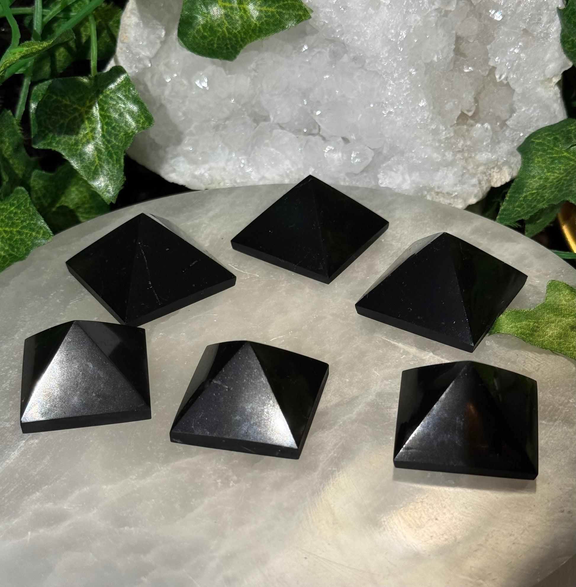 Black Shungite Pyramid  on a stone surface with a crystal and greenery in the background